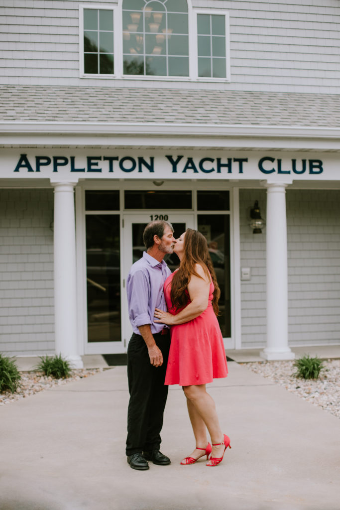 Couple kissing in front of their wedding venue, the Appleton Yacht Club, during their engagement session.