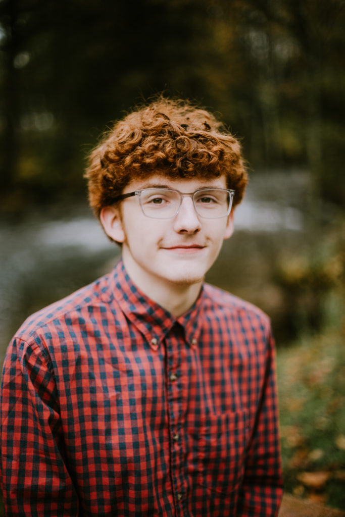 Curly haired red headed boy in glasses poses for his senior pictures in a red plaid button down shirt. Class of 2023 at Waupaca High School.