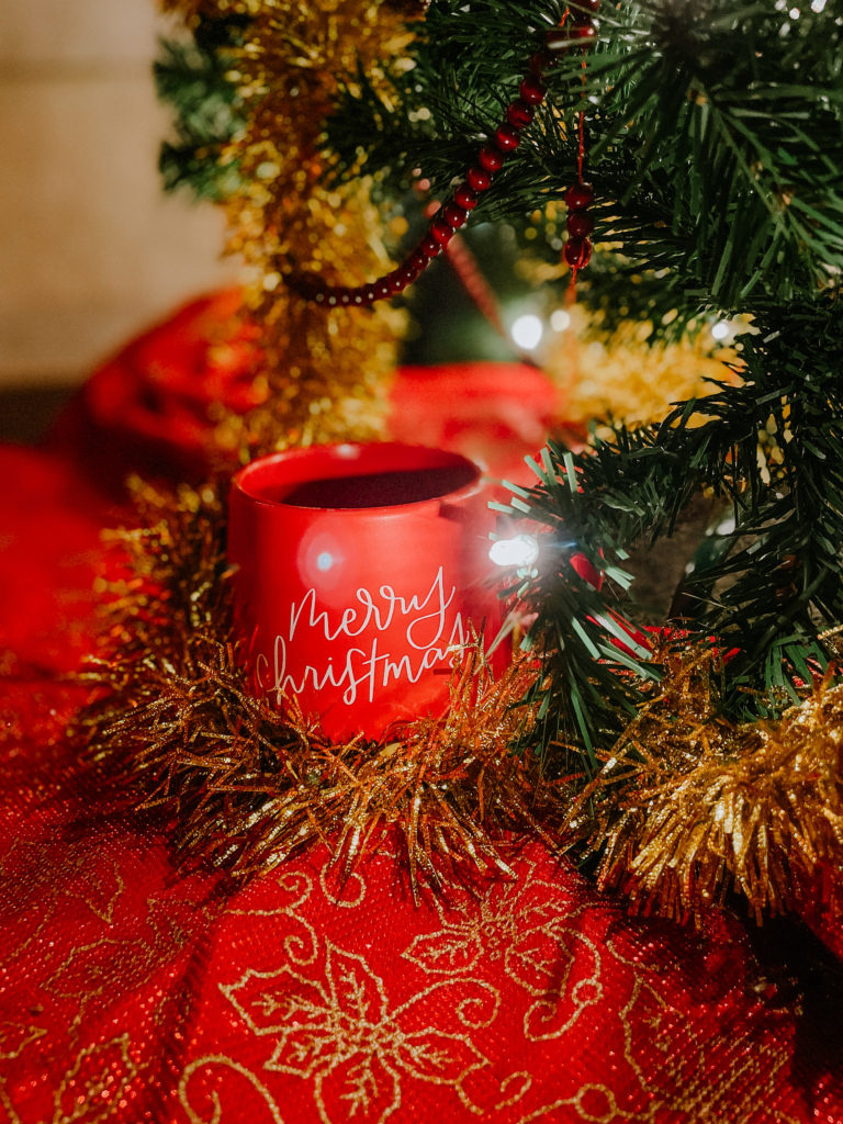 Red Merry Christmas mug sitting on a red and gold tree skirt in front of a green Christmas tree decorated with red beads and gold tinsel. Currently at Anna Gutermuth Photography.