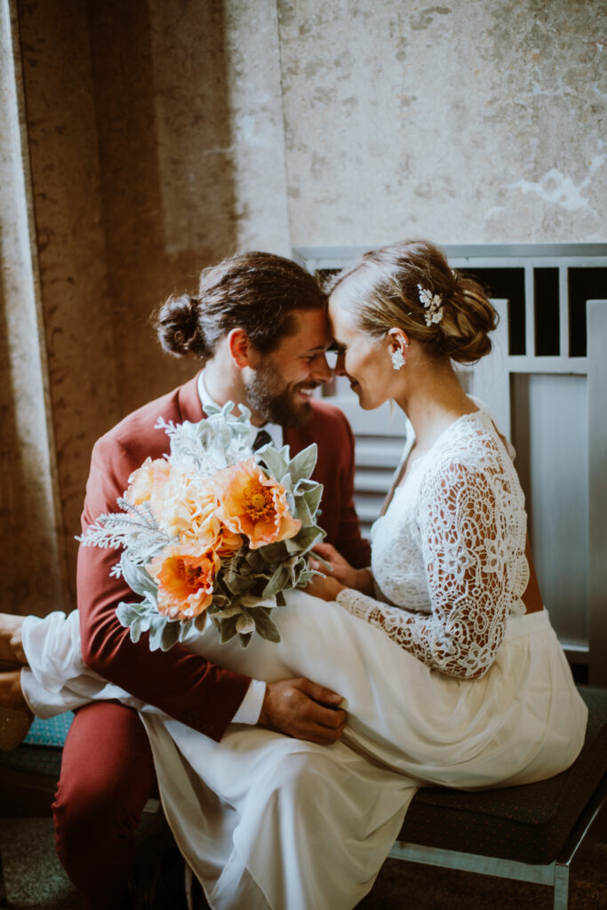 Bride in a lacy white dress holding her orange bouquet and groom in a red suit sitting on a bench surrounded by 1940s marble architecture during their courthouse elopement.