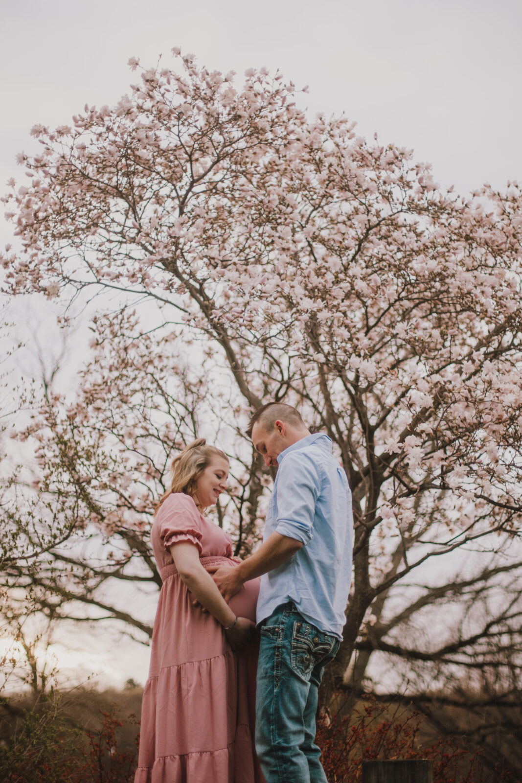 Pregnant woman in a pink dress is holding her belly and cradled by her husband in a blue button down shirt and jeans during their Green Bay maternity session