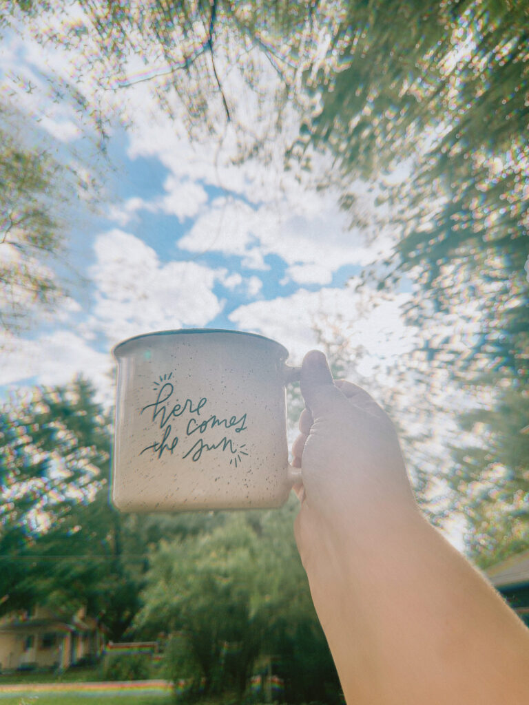 Coffee mug reading here comes the sun being held up in the air by a hand in front of green trees and blue sky. Currently, behind the scenes at Anna Gutermuth Photography.