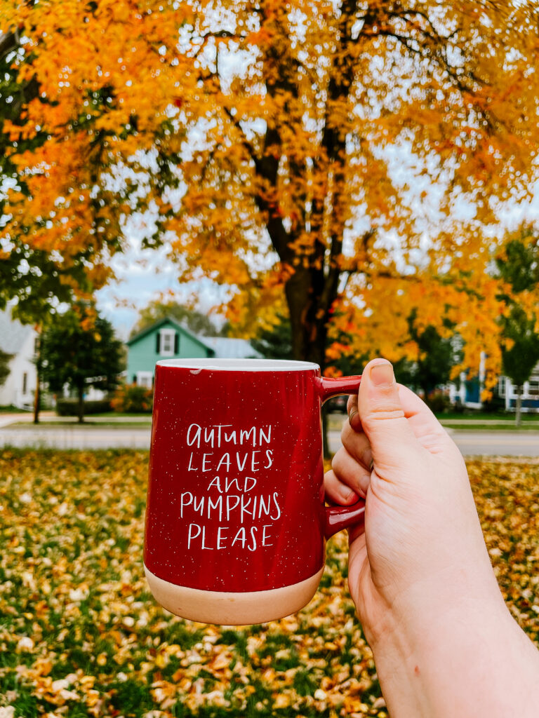 Currently... Red coffee mug that reads Autumn Leaves and Pumpkins Please in front of a tree with orange leaves