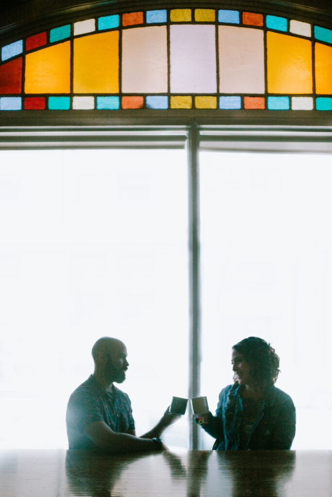 Couple cheersing with lattes in front of a big window with stained glass during their coffeehouse engagement session