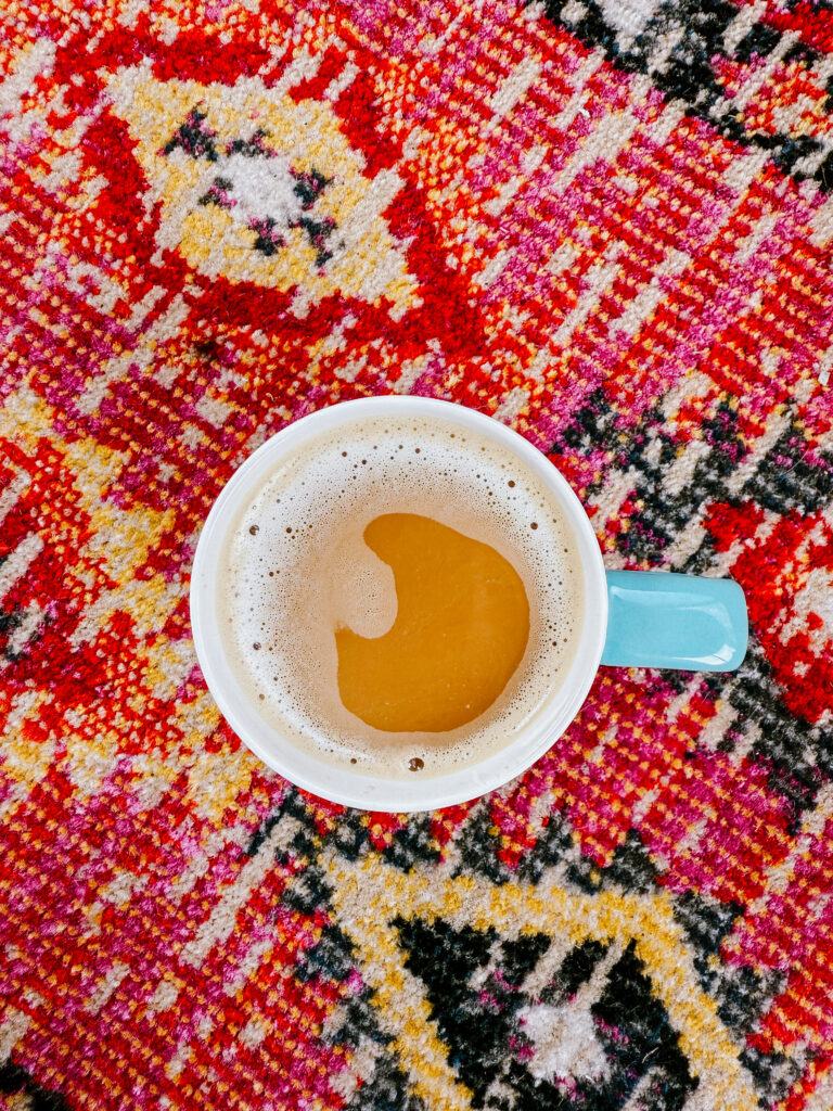 Currently drinking coffee out of a blue mug sitting on the porch with a red pink yellow and black tribal print rug.